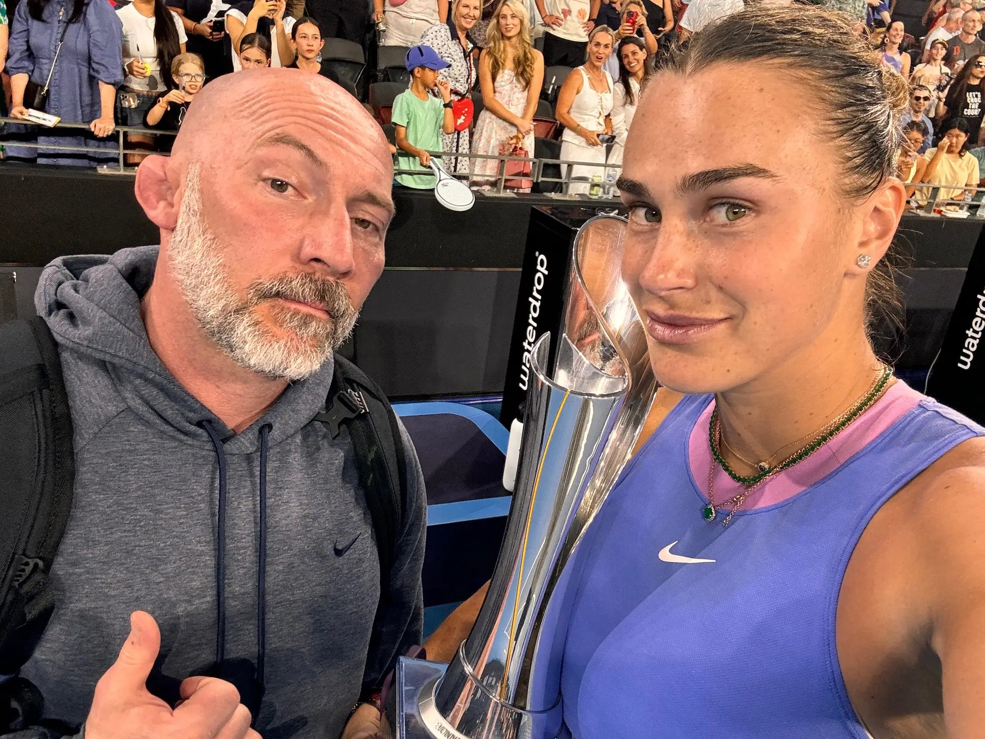 Jason Stacy with Aryna Sabalenka holding a tournament trophy courtside